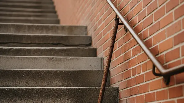 A steep outdoor concrete staircase with a rusted, detached handrail leaning against a brick wall.