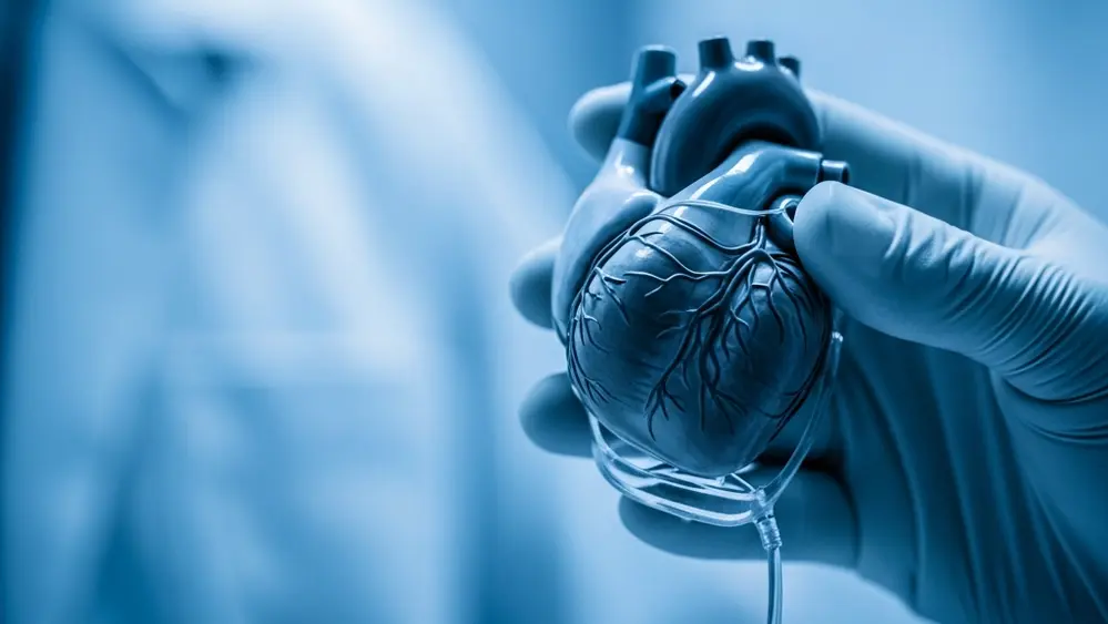 A close-up of a sophisticated medical heart device held by a gloved hand in a sterile hospital setting with soft blue lighting.