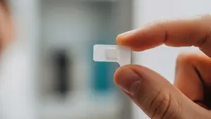 A close-up view of a small, translucent medicinal film strip being held between two fingers, representing a Suboxone sublingual film, with a soft-focus background of a bathroom medicine cabinet.