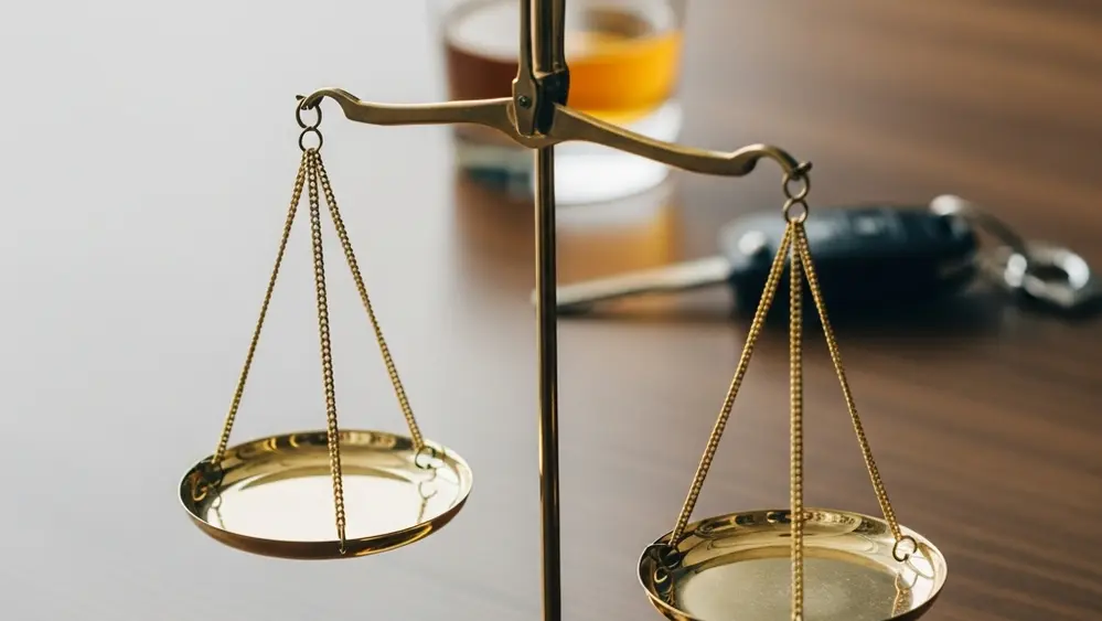 A symbolic image showing a set of gold scales of justice on a wooden desk, with a set of car keys and a partially empty glass of amber liquid sitting nearby in a blurred background.