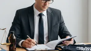 A professional person in business attire sitting at a clean wooden desk, holding a pen and reviewing a large set of papers with a focused expression.