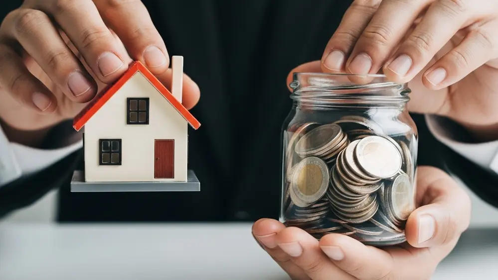 A close-up view of a person's hands holding a miniature model of a house and a small jar of coins, representing retirement savings and financial security.