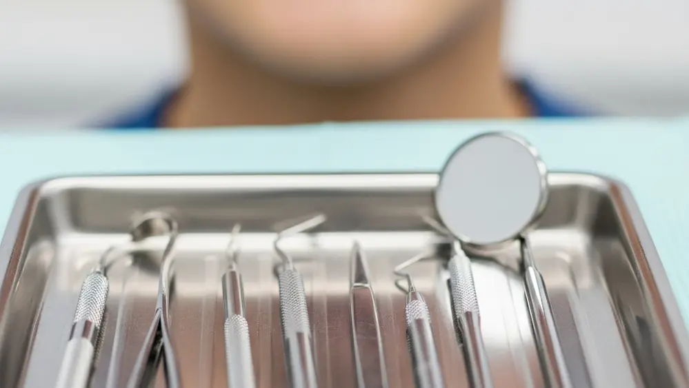 A close-up of a dentist's tray with various metal instruments and a dental mirror, with a person's chin and mouth visible in the blurred background.