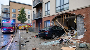 An apartment building with a large hole in a ground floor unit where a dark sedan has crashed through the wall, surrounded by debris and emergency vehicle lights.