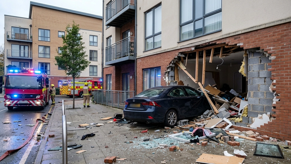 An apartment building with a large hole in a ground floor unit where a dark sedan has crashed through the wall, surrounded by debris and emergency vehicle lights.