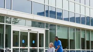 A modern hospital exterior with glass windows and a patient being assisted by a healthcare worker in a wheelchair near the entrance.