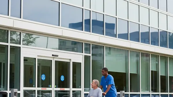 A modern hospital exterior with glass windows and a patient being assisted by a healthcare worker in a wheelchair near the entrance.