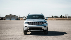 A modern silver Jeep SUV parked alone in a large empty gravel lot during the daytime, with a blurred residential garage in the distant background.