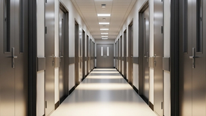 A perspective view of a long institutional hallway with metallic doors and shadows cast from ceiling lights.