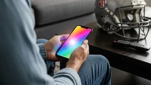 A person sitting on a sofa holding a smartphone that glows with colorful interface elements, while an NFL football helmet and a remote control sit on a nearby coffee table.