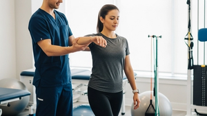 A physical therapist wearing a navy blue uniform assisting a female patient with balance exercises in a bright clinical setting with exercise equipment.