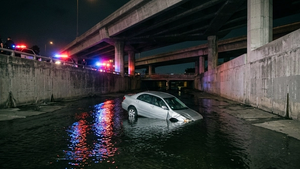 A silver sedan submerged in a concrete canal under a freeway overpass with emergency lights reflecting on the water surface.