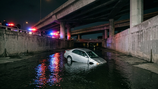 A silver sedan submerged in a concrete canal under a freeway overpass with emergency lights reflecting on the water surface.