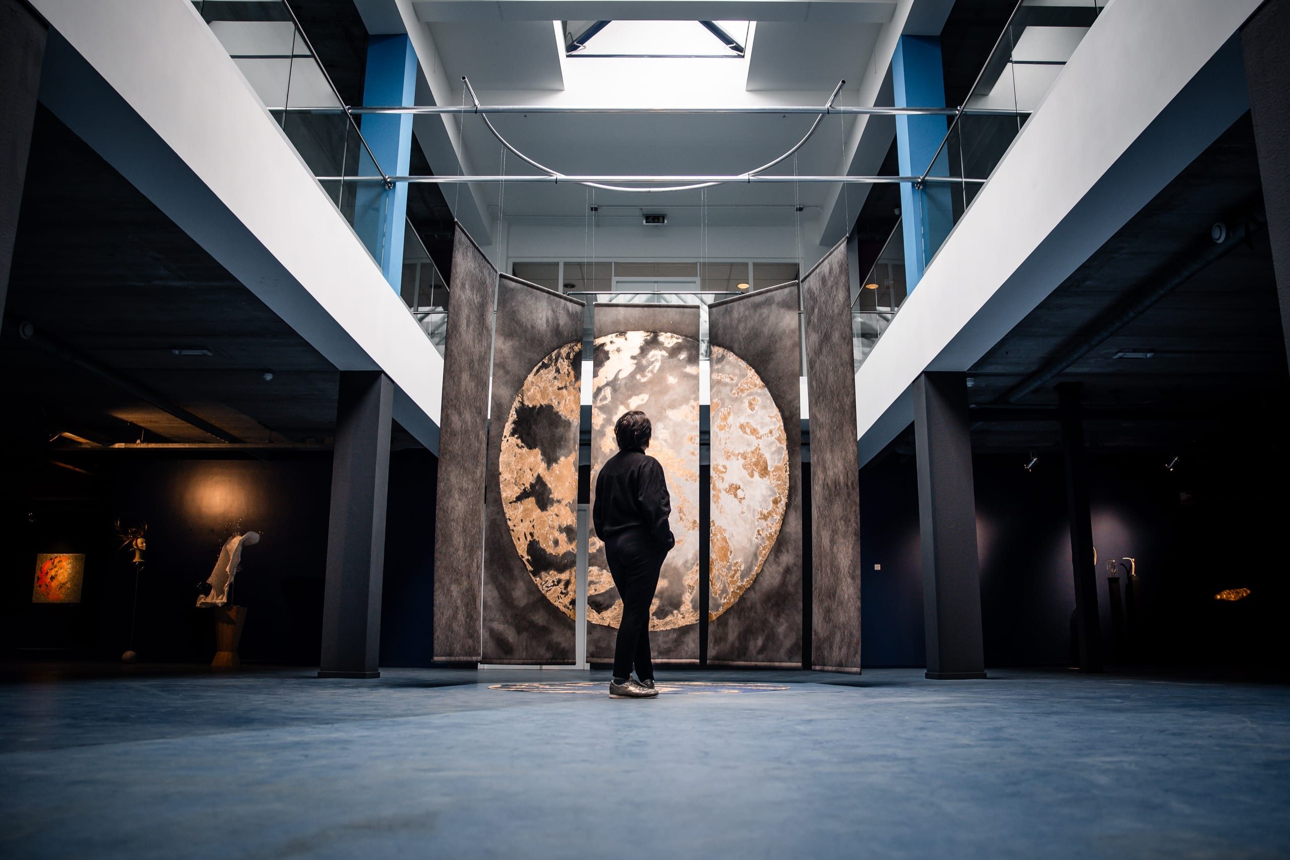 A visitor stands in front of Connected: five hanging panels that together form a moon in gold and grey.