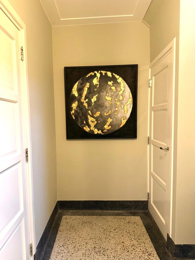 Een ronde maan in grijs en goud in een zwarte lijst op de kopse wand van een hal met terrazzovloer en panelen deuren.