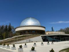 The Silesian Planetarium from the forecourt: the silver dome on its round stone ring, a glass pavilion set into the slope below it, and young pines in concrete planters across the square.