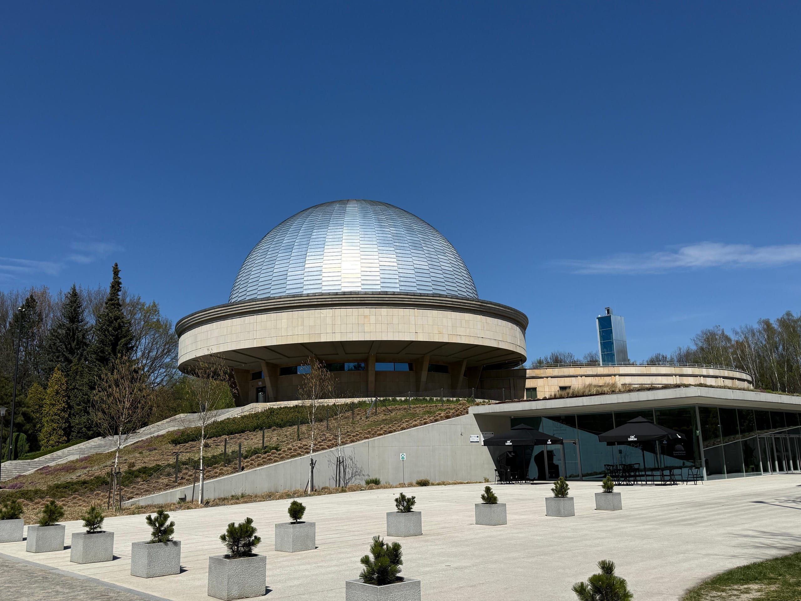 The Silesian Planetarium from the forecourt: the silver dome on its round stone ring, a glass pavilion set into the slope below it, and young pines in concrete planters across the square.