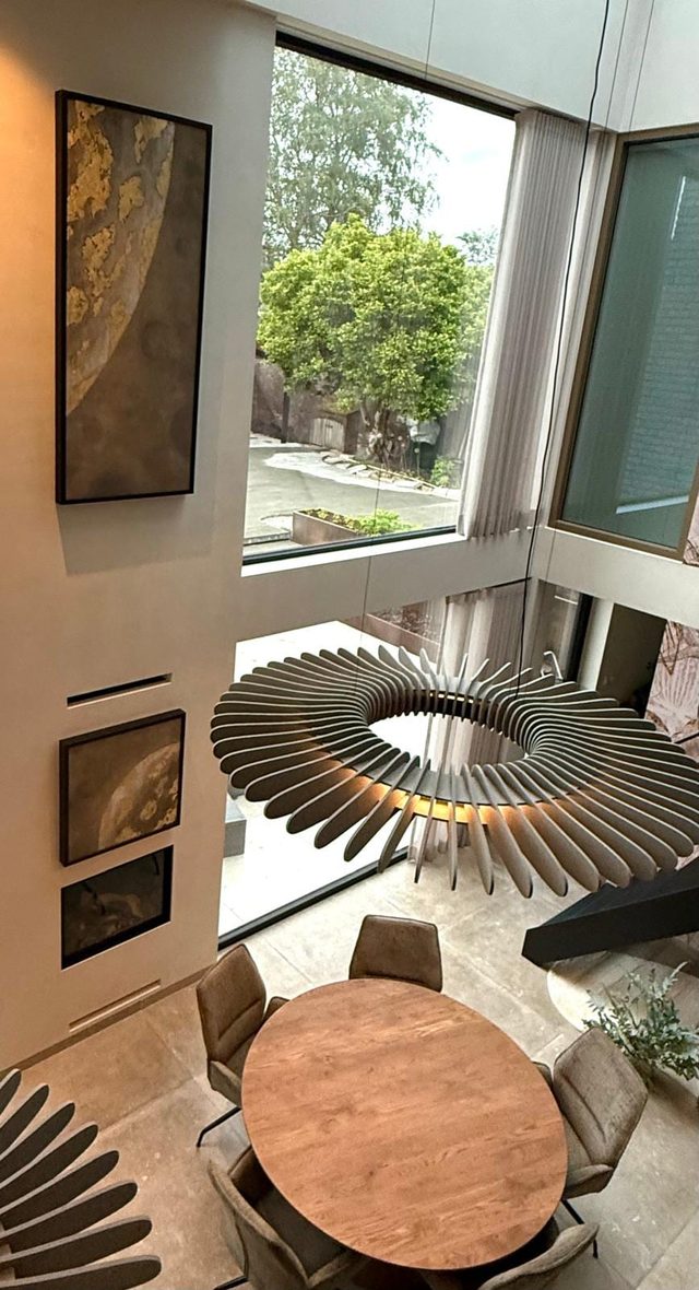 Een vide van twee verdiepingen met twee planeetschilderijen op de wand, een ronde eettafel eronder en glas over de volle hoogte naar de tuin.