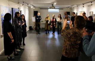 Visitors in the room during the opening, blue and gold planetary paintings along both walls and someone addressing the gathering.