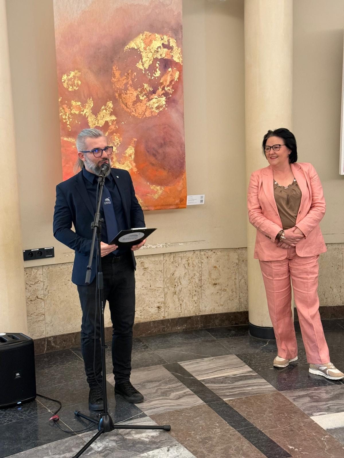 A man wearing a planetarium lanyard speaks into a microphone from a folder, in front of the tall coral work with gold-coloured metal leaf; a visitor listens beside him.