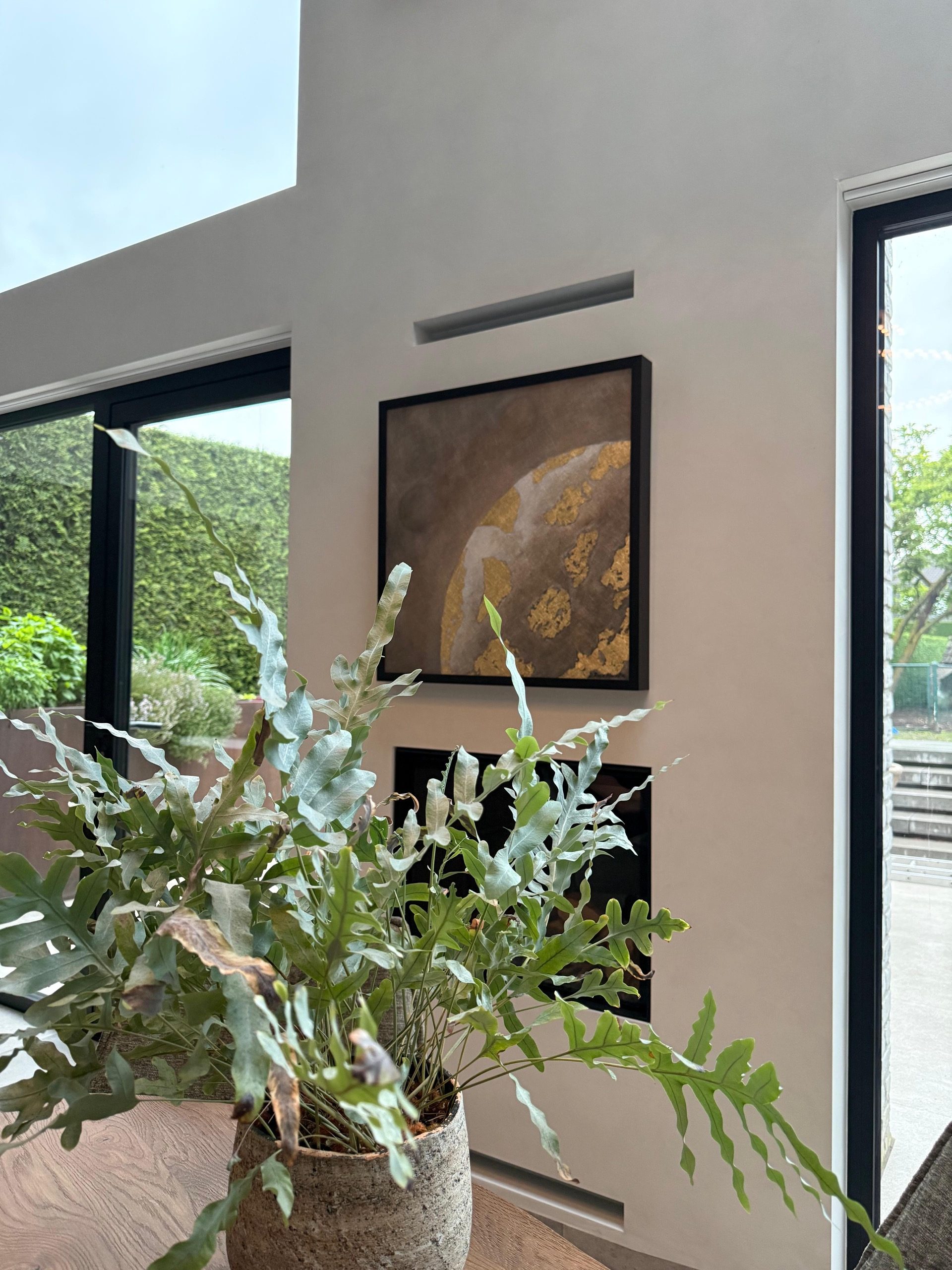 A square planetary painting in brown and gold above a clean built-in fireplace, between two glass walls onto the garden.
