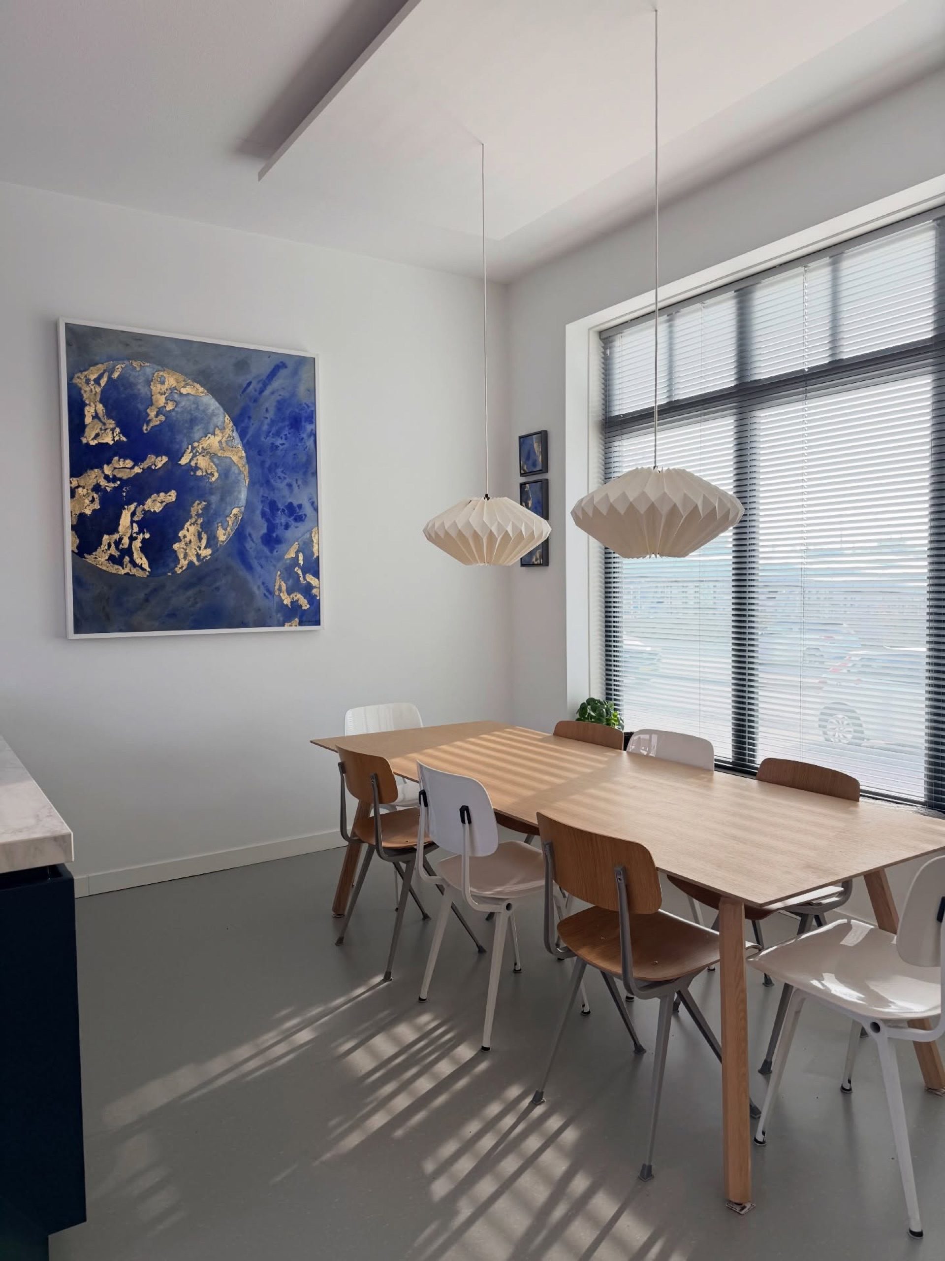 A large blue and gold planetary painting on the wall beside a long dining table, in a bright interior with a tall window.