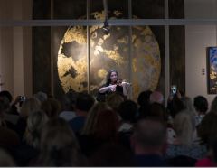 A violinist plays in front of the lit work, the audience seen from behind.