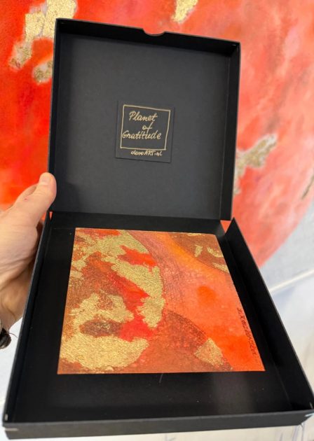 A small square work in orange and red with gold-coloured metal leaf, in a black box.