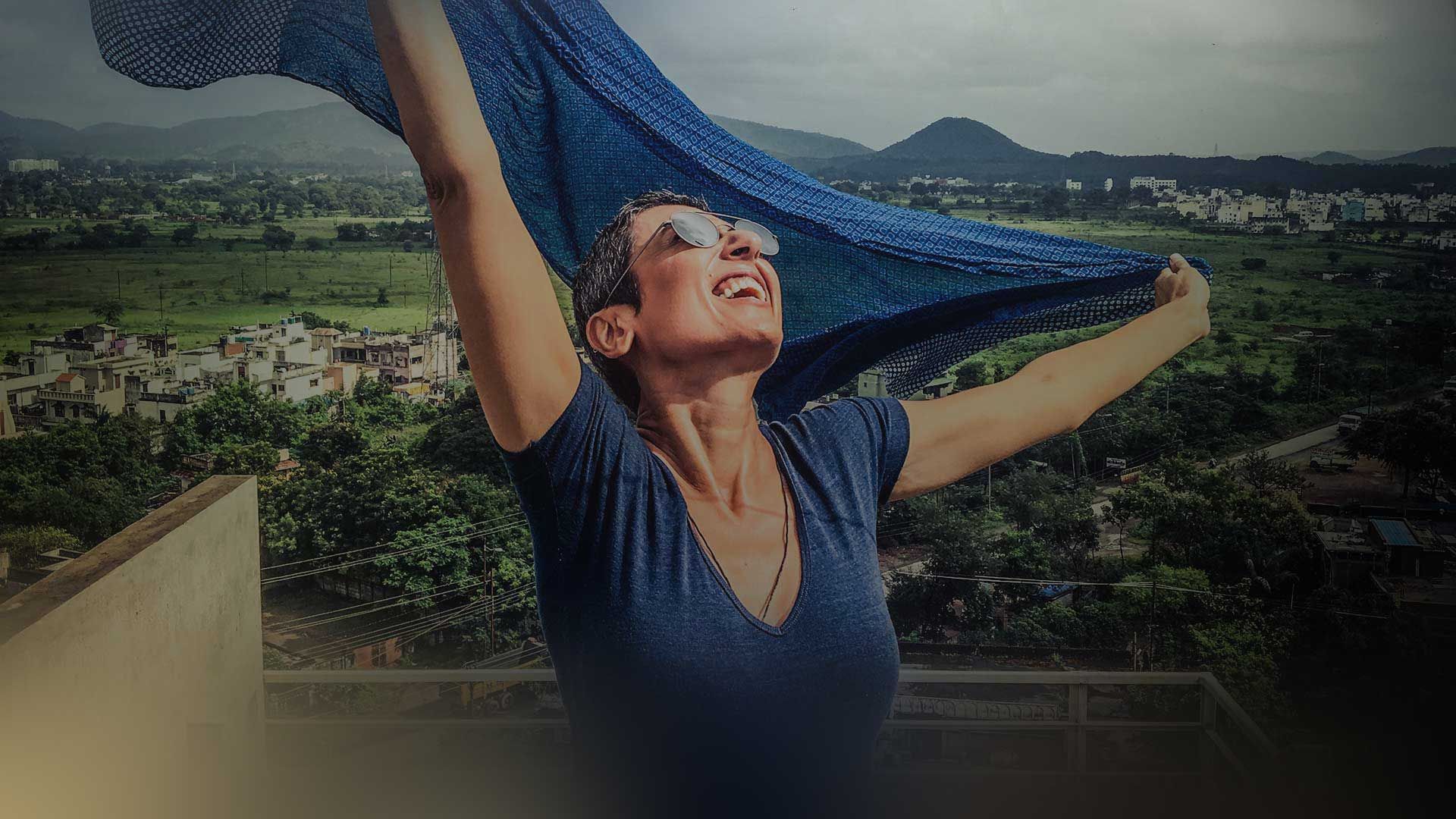 Zainab Salbi on a rooftop, arms raised, a blue scarf lifting in the wind