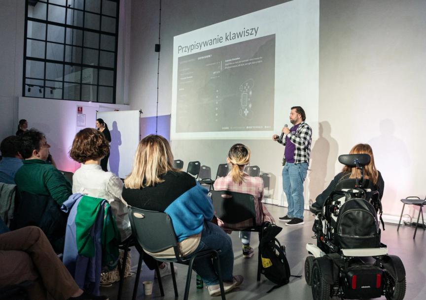 A man - Mateusz Pusty stands in front of a crowd with a microphone in his hand and talks about accessibility. Behind his back there's a big screen on the wall.