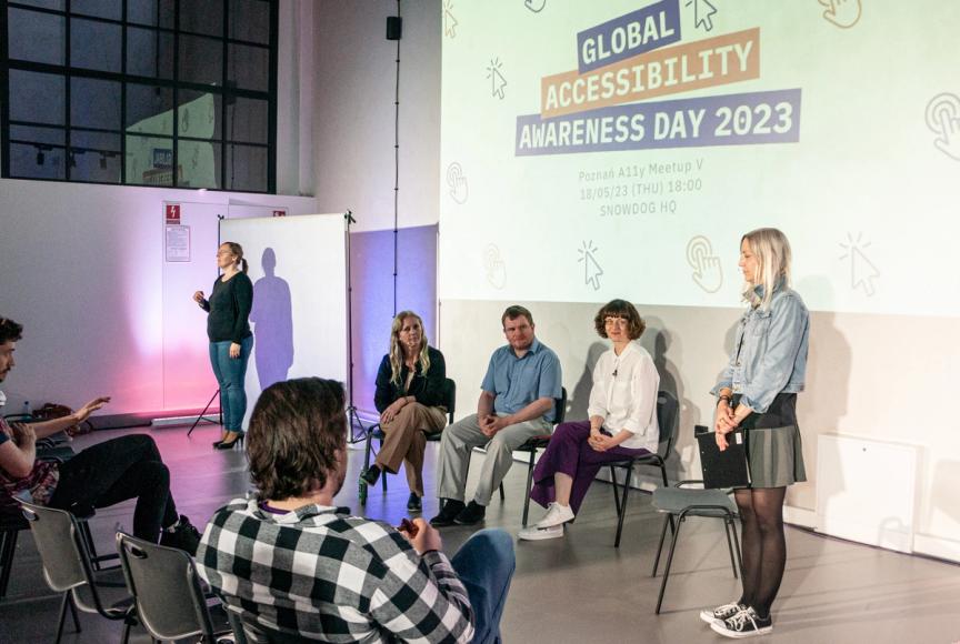 Global Accessibility Awareness Day 2023. In front of an audience there are 3 people sitting in chairs. A blonde woman, a man and a brown haired woman. Right next to them stands Ania Karoń - blond woman who's a moderator of the conversation. On the left side there's a sign language translator.