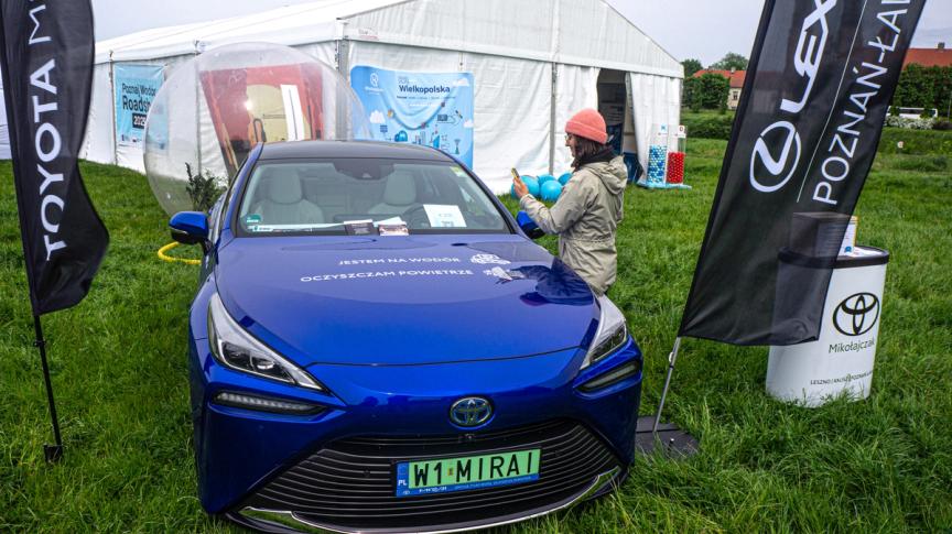 Samochód wodorowy Toyota Mirai stojący przed namiotem ekspozycji Poznaj Wodór Roadshow. Uczestniczka badania sczytuje kod QR znajdujący się na samochodzie. 
