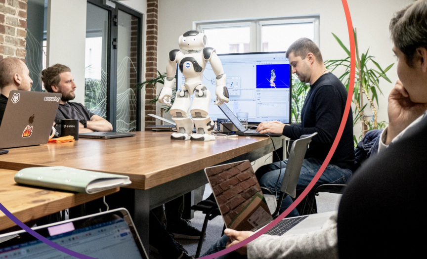 NAO the robot standing on a desk surrounded by a group of people studying the Choreographe software displayed on the screen behind the robot.