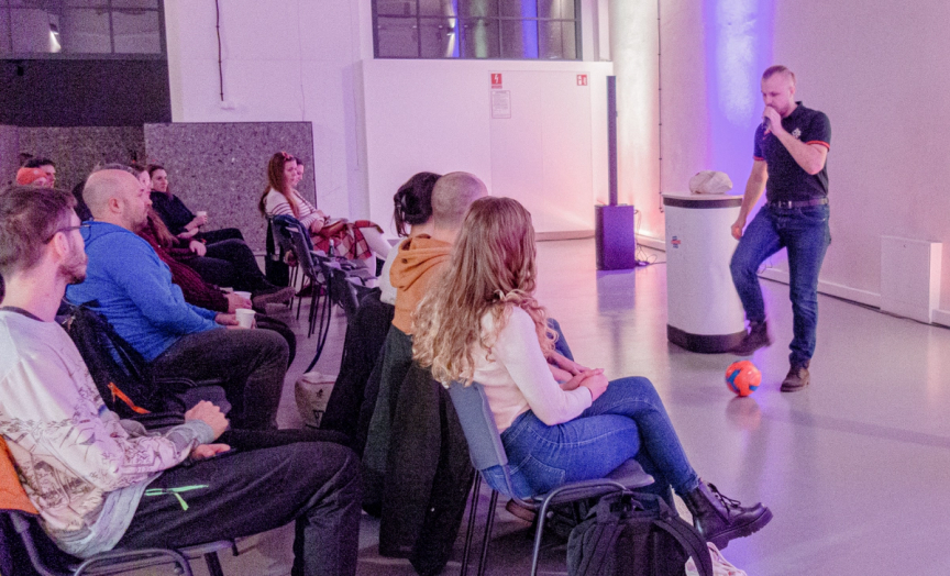 On the left side you can see a group of people, sitting in charis, who came to the meetup. On the right hand side there’s Grzegorz Jakubowski. The first speaker at the Poznan Accessibility Meetup 3. He is standing with microphone in his hand and a special football by his foot.