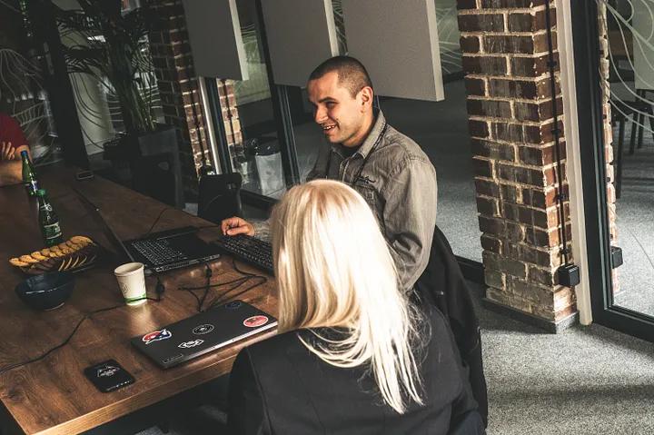 Two people sitting by the desk. The man — Przemek, is in front of the computer testing software. The woman — Ania, is sitting and listening to him speaking.