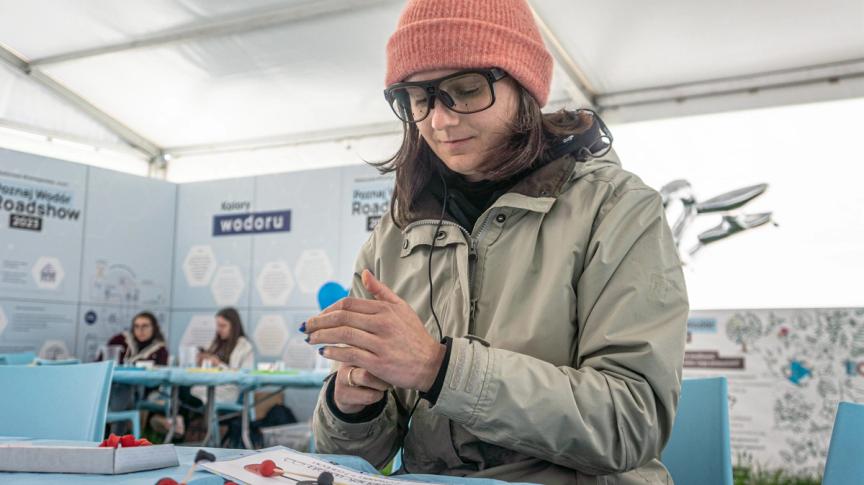 Kobieta siedzi przy stole i lepi cząsteczki z plasteliny. Widoczne okulary do eye trackingu z punktowymi kamerami zatopionymi w szkłach.