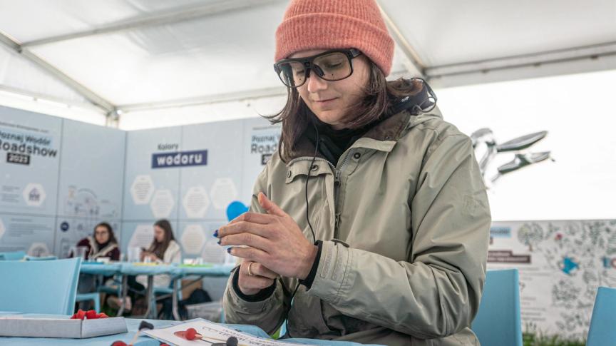 Kobieta siedzi przy stole i lepi cząsteczki z plasteliny. Widoczne okulary do eye trackingu z punktowymi kamerami zatopionymi w szkłach. 