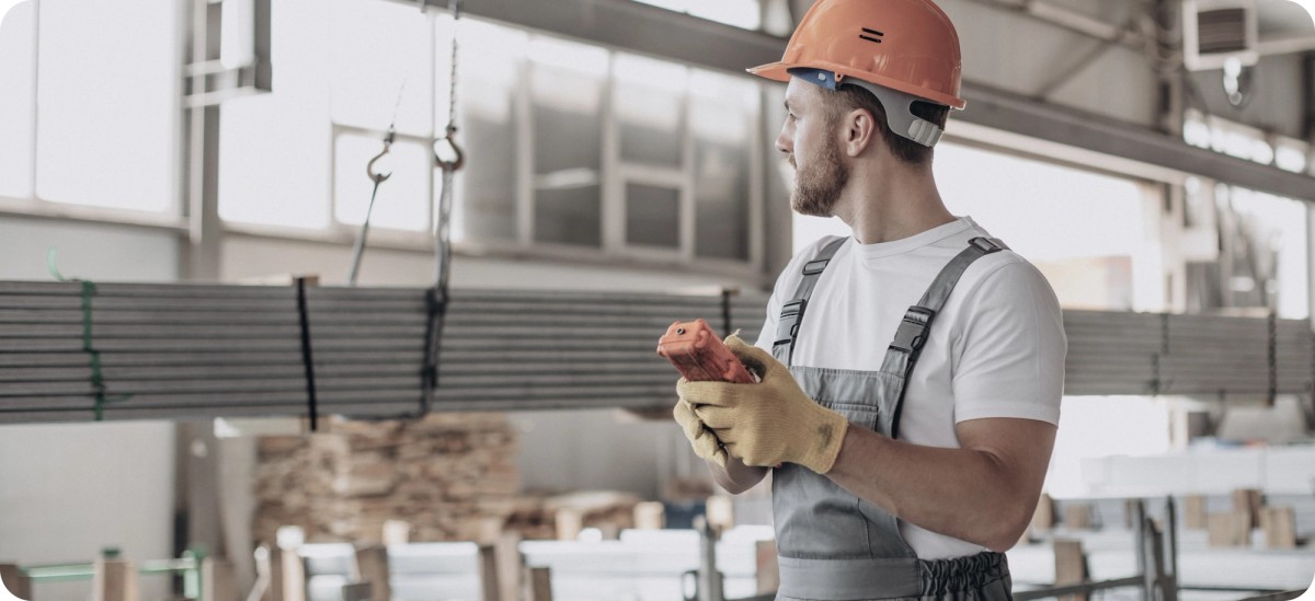 Работник със защитна каска и ръкавици контролира транспортиране на строителни материали в производствено хале – илюстрация за приложението на Цифров продуктов паспорт в строителния сектор.