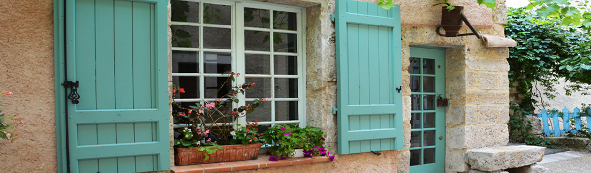 A photo of a house in France with flowers in the window ledges. The doors and windows of the property are painted in a teal blue. 