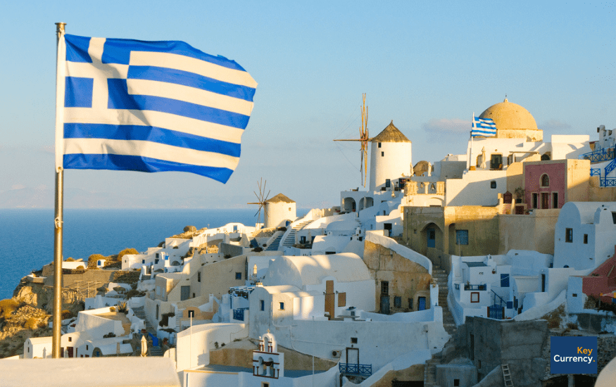 Greek flag waving in the wind with a backdrop of Santorini, a city in Greece on a sunny day.
