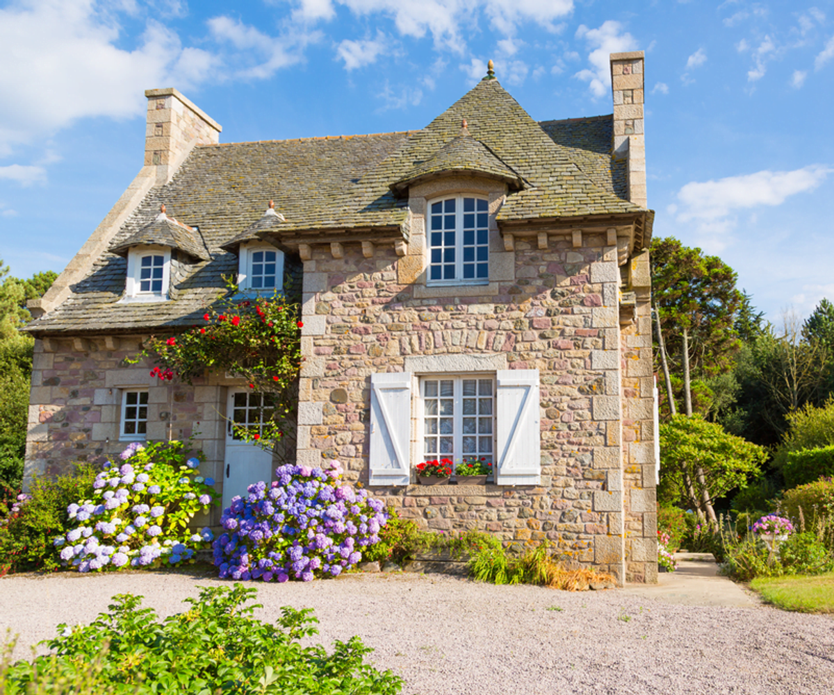 Photo of a French property showing the whole house. Various colourful flowers are in front of the property that are both pink, red and lavender. 