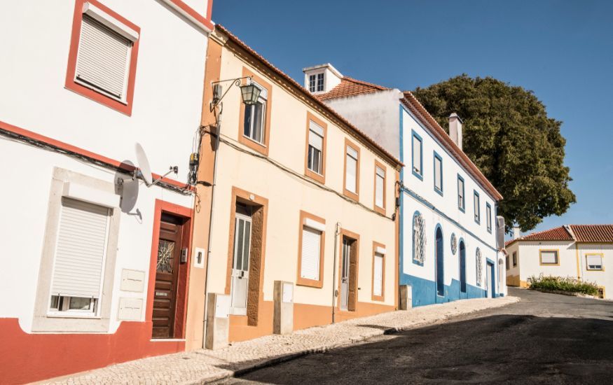 Photo of a street in Portugal and different properties on the street. Blue skies and large tree in the background of the photo.