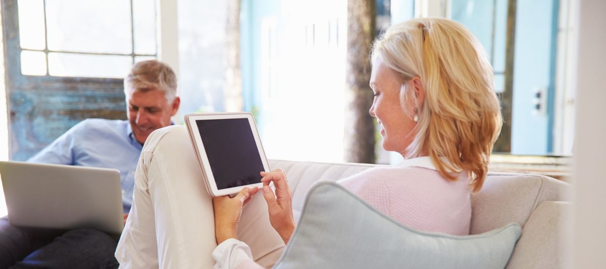 A women using a tablet and a man using a laptop both sat on a sofa in their home. 