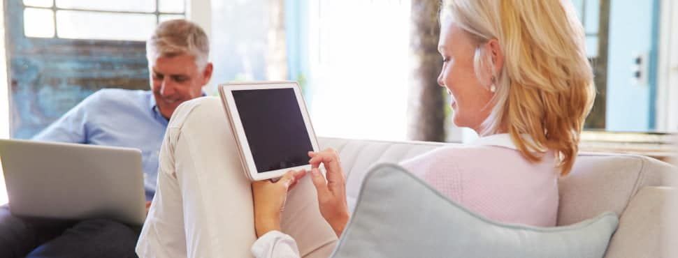 A women with a tablet sat on one end of the sofa at home with a man using a laptop on the other side.