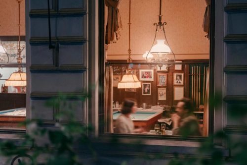 Blick von der Straße durchs Fenster ins abendliche Billardzimmer, ein Paar im Gespräch