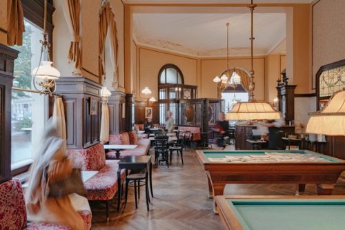 Billardtisch mit ausgelegten Zeitungen, dahinter der Saal mit Gästen und Kellnern in Bewegung