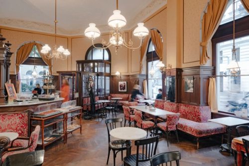 Ecke des Kaffeehauses mit Lustern, Mehlspeisvitrinen, roten Polsterbänken und Bugholzstühlen