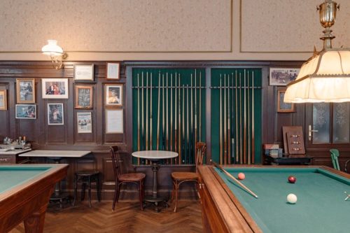 Billardqueues an grüner Wand, davor ein Billardtisch mit drei Kugeln und gerahmte Fotos an der Holzvertäfelung