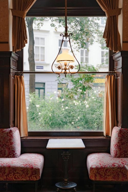 Marmortisch zwischen zwei roten Polsterbänken am Fenster, darüber eine Hängelampe, draußen blühende Rosen