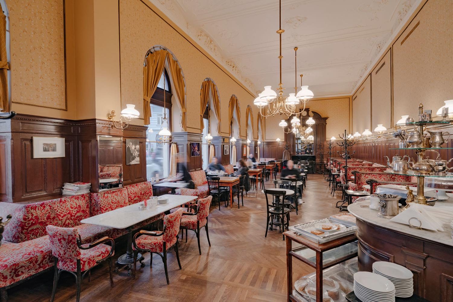 Langer Saal des Café Sperl mit Rundbogenfenstern, Messinglustern, roten Polsterbänken und Marmortischen